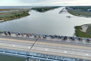 drone shot of large group of riders crossing a bridge over a river with marsh and houses in the background